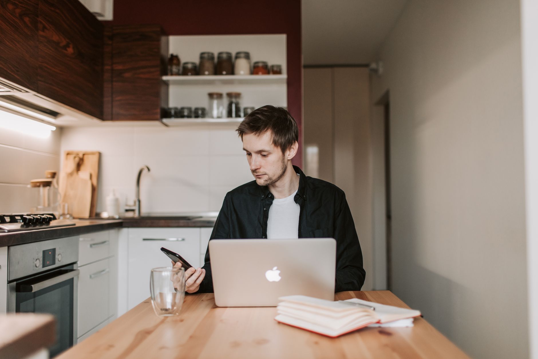 thoughtful male freelancer using smartphone and laptop in kitchen