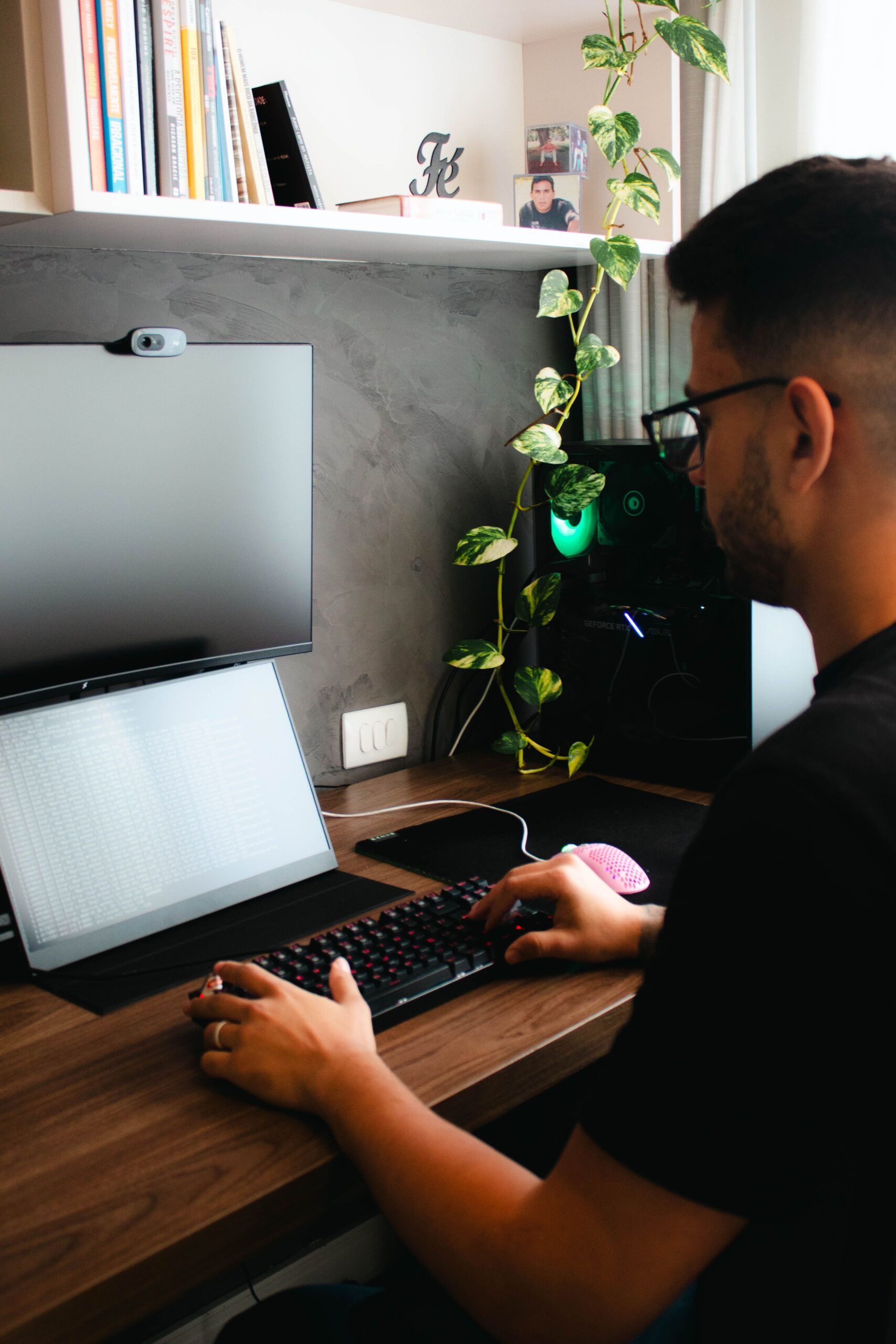 Person using a laptop beside a monitor and plants.