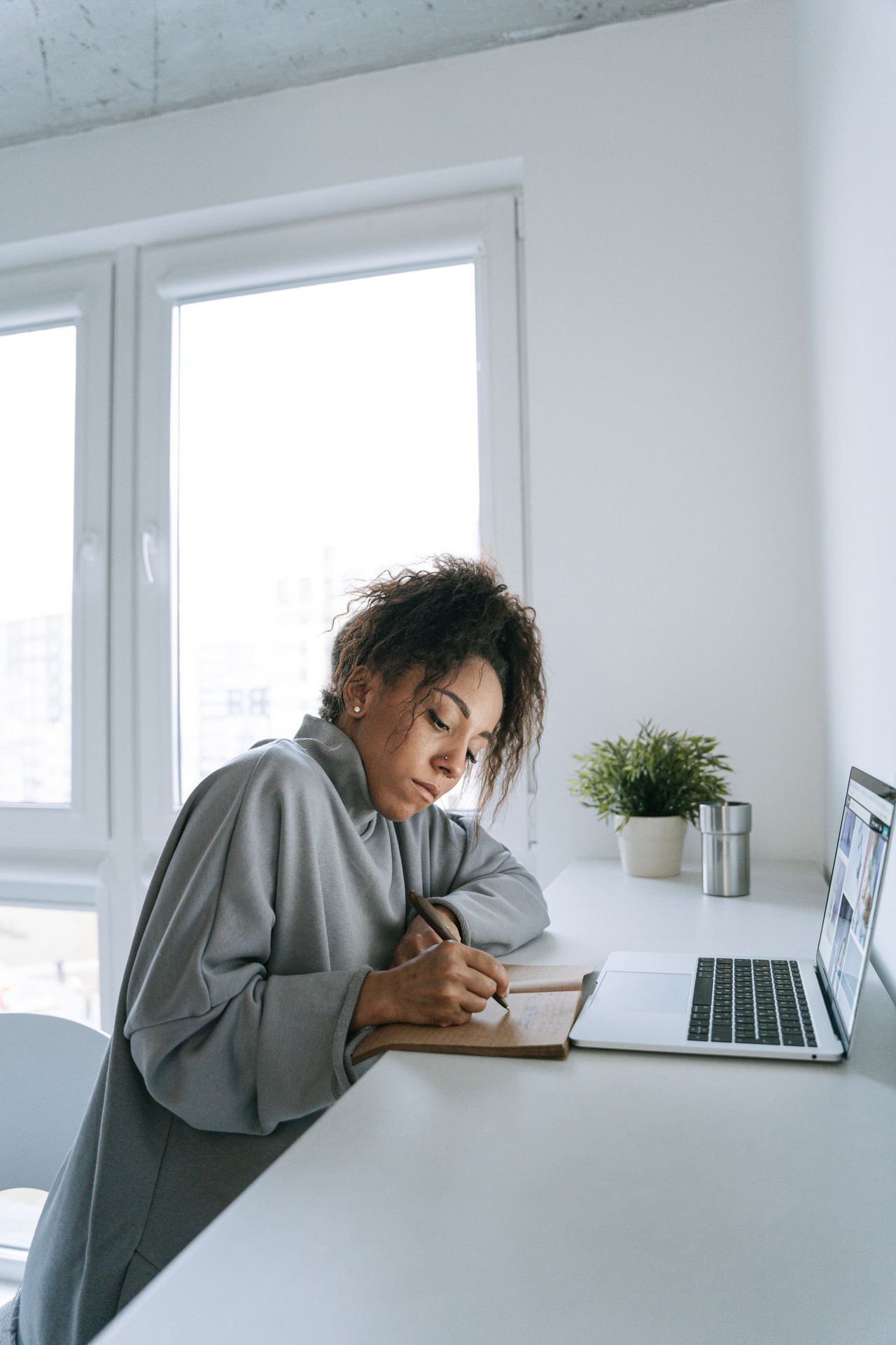 Person writing in a notebook beside a laptop near a window.