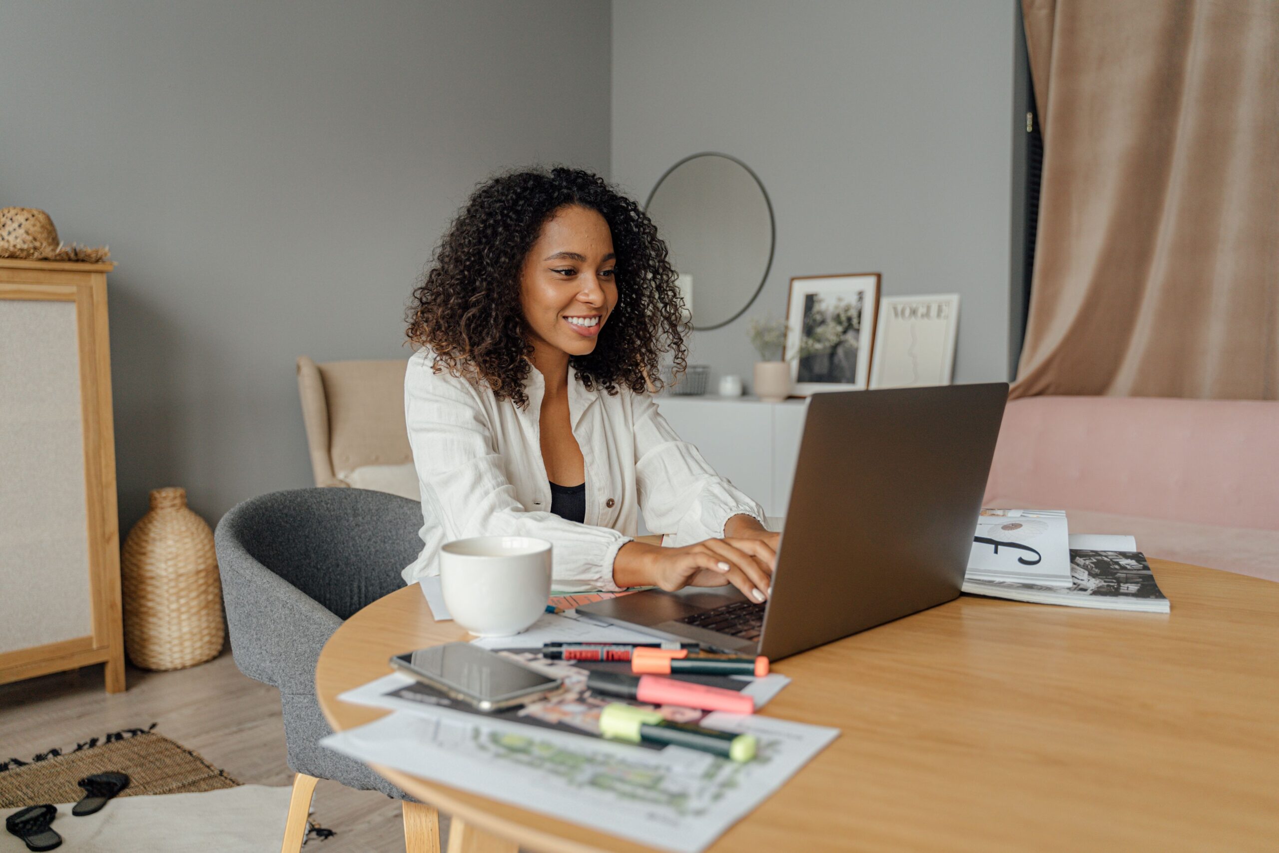 Smiling person using a laptop at a table with notebooks and a mug.
