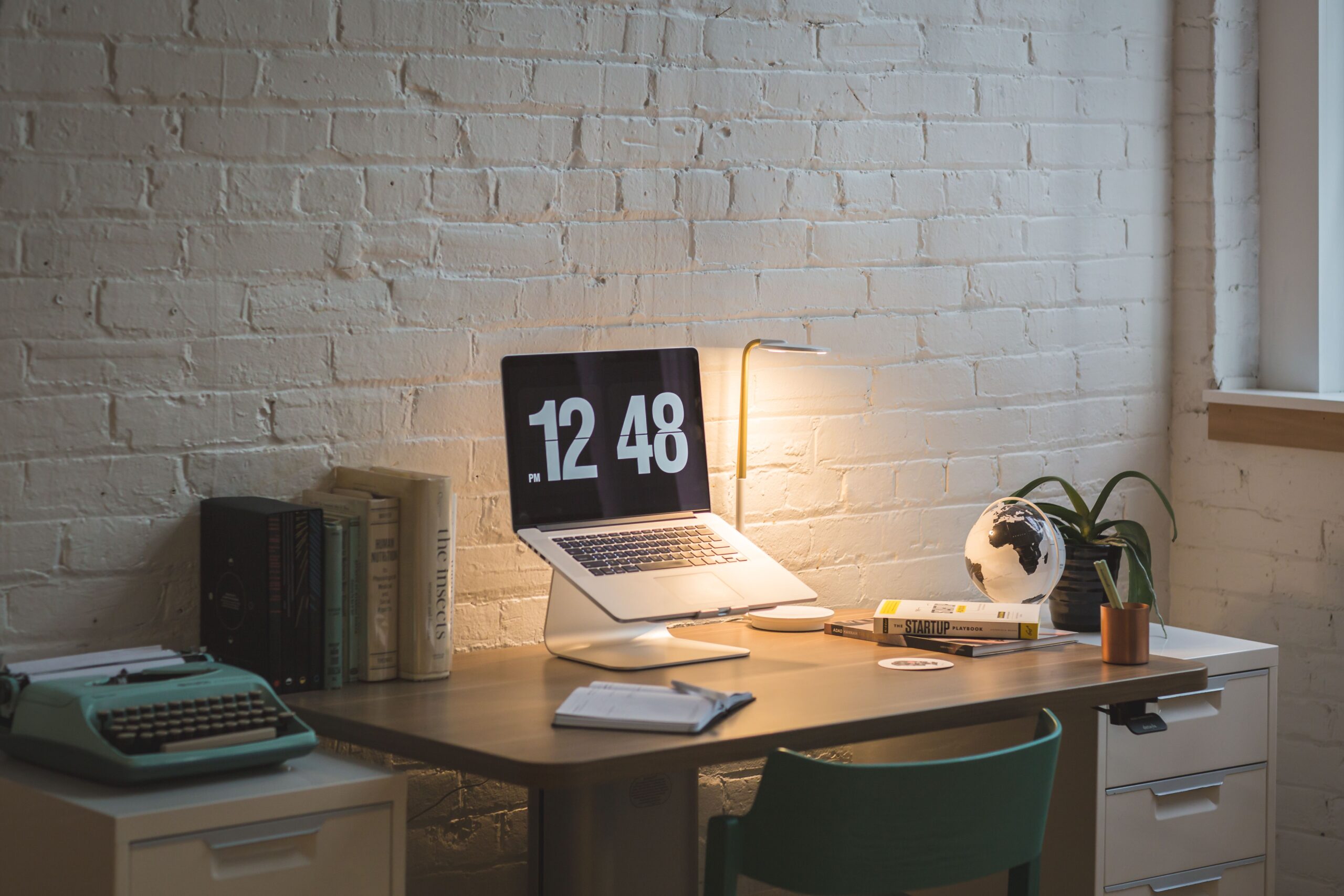 Laptop, books, desk lamp and chair beside a brick wall.