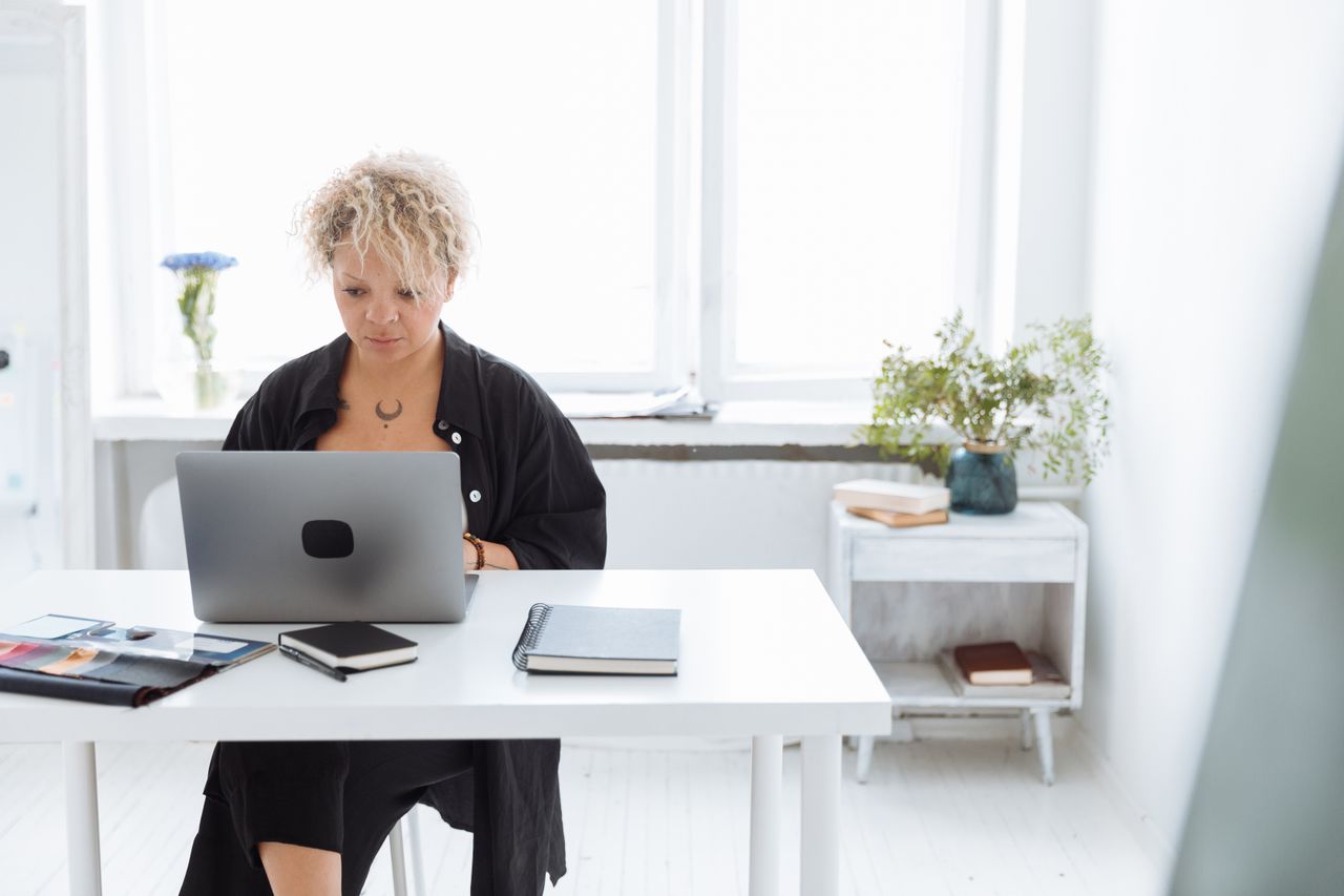 Person at a desk with a laptop, notebook and plants beside a window.