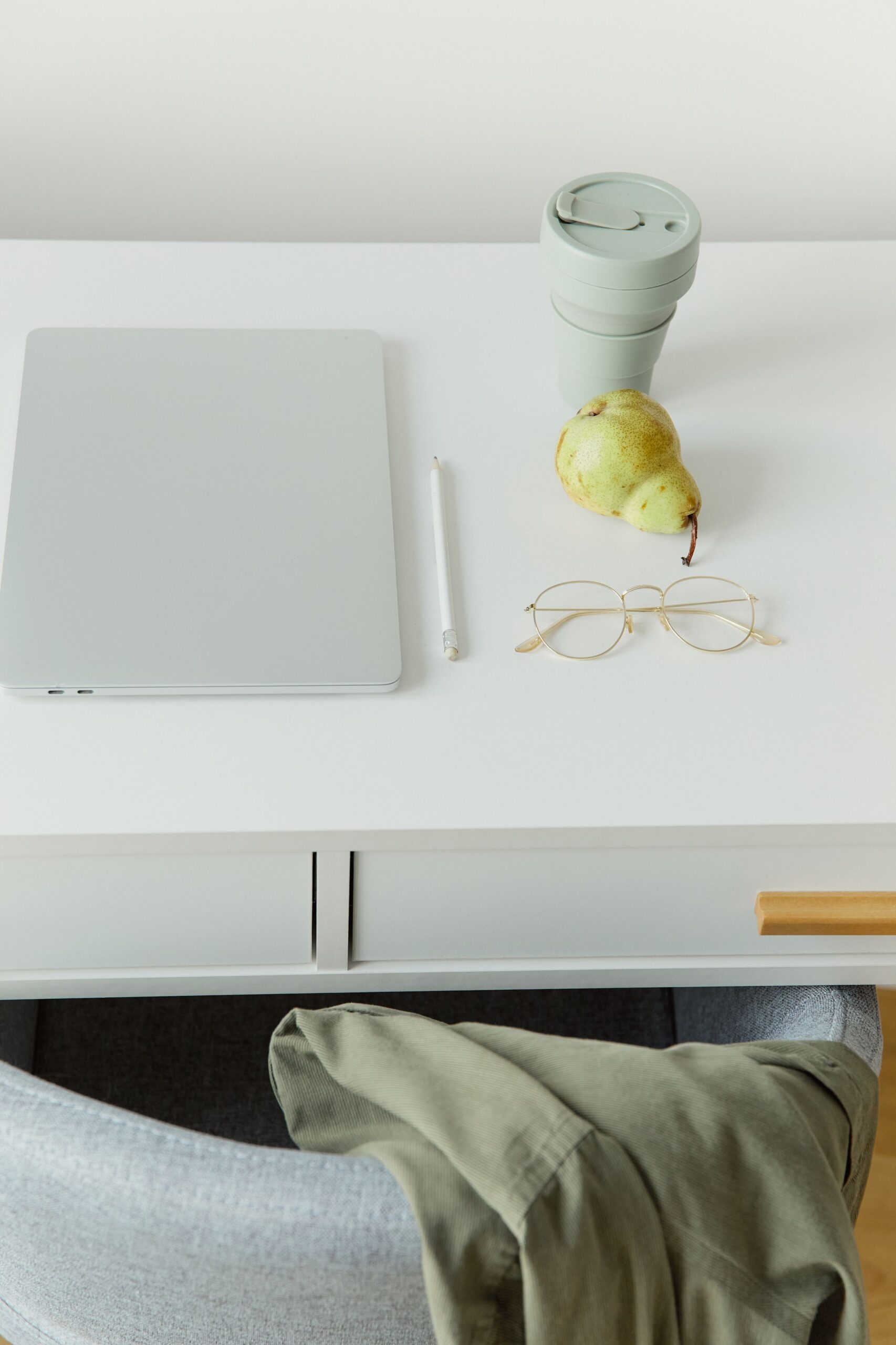 Laptop, eyeglasses, cup and pear on a white table.
